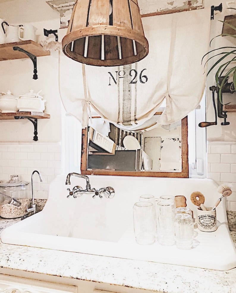 May include: A white farmhouse style kitchen sink with a chrome faucet and a wooden basket hanging above. The sink has a white countertop and a white tiled backsplash. There are several glass jars and a glass pitcher on the counter. The basket has the number "26" printed on it.