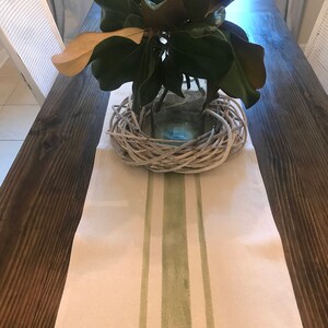 May include: A white table runner with two green stripes down the center. The runner is on a dark brown wooden table with a glass vase filled with green leaves and a white woven wreath.