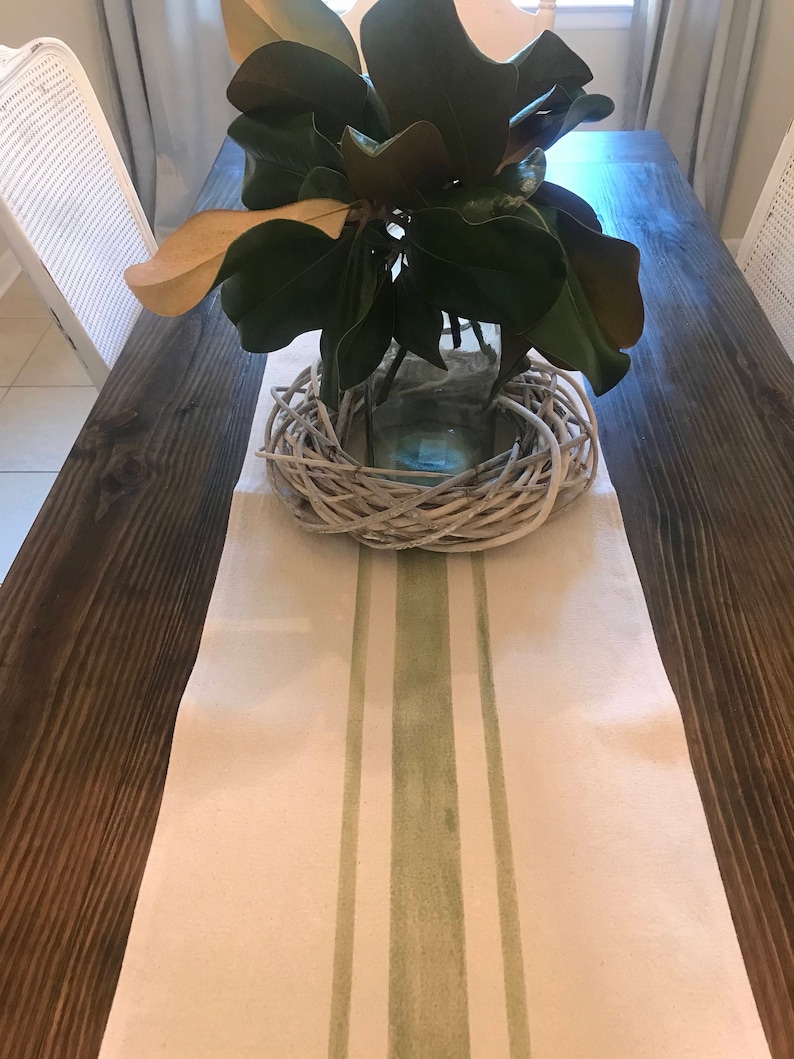 May include: A white table runner with two green stripes down the center. The runner is on a dark brown wooden table with a glass vase filled with green leaves and a white woven wreath.