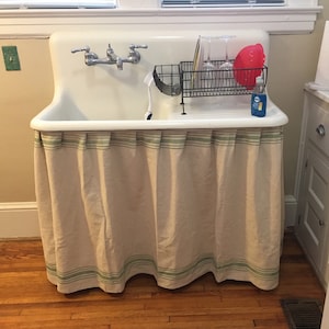 May include: A white porcelain kitchen sink with a chrome faucet and a beige fabric skirt with green stripes. The sink has a dish rack with glasses and a red bowl on top.