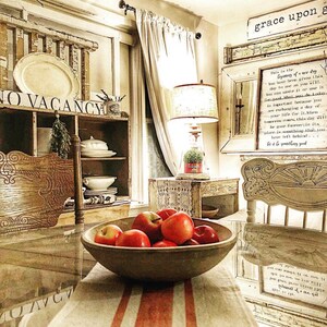 May include: A rustic farmhouse kitchen with a wooden table set with a bowl of red apples. The table has a white and red striped tablecloth. The kitchen features a vintage cabinet with glass doors, a lamp with a white shade, and a sign that reads "grace upon grace".