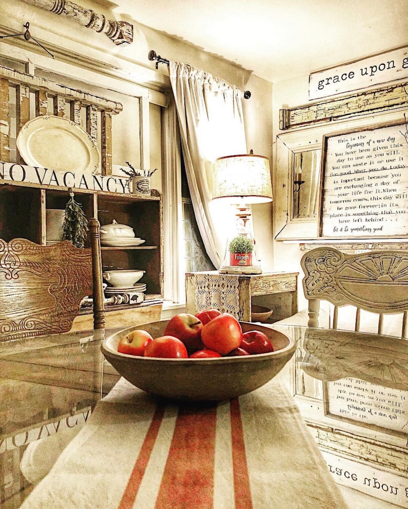 May include: A rustic farmhouse kitchen with a wooden table set with a bowl of red apples. The table has a white and red striped tablecloth. The kitchen features a vintage cabinet with glass doors, a lamp with a white shade, and a sign that reads "grace upon grace".