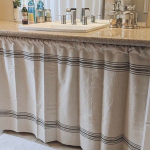 May include: A white bathroom vanity with a beige and brown striped fabric curtain covering the lower portion of the cabinet. The vanity has a white countertop with a white porcelain sink and chrome faucets.