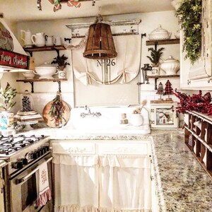 May include: A white farmhouse kitchen sink with a white countertop and white cabinets. The cabinets have a white ruffle trim. The sink is decorated with Christmas decorations, including a small Christmas tree, a wreath, and a red and white candy cane.