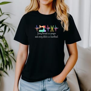 May include: Black t-shirt featuring a colorful sewing machine, scissors, and heartbeat design. The text reads, "Every thread is a prayer and every stitch is a heartbeat." A casual, everyday garment.