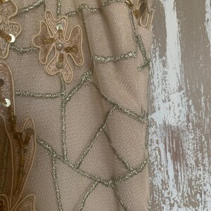 May include: Close-up of a beige fabric with intricate detailing. The fabric features a net-like pattern with silver thread accents. Floral and geometric designs are embellished with sequins and beads, creating a textured and elegant appearance. The overall aesthetic suggests formal wear.