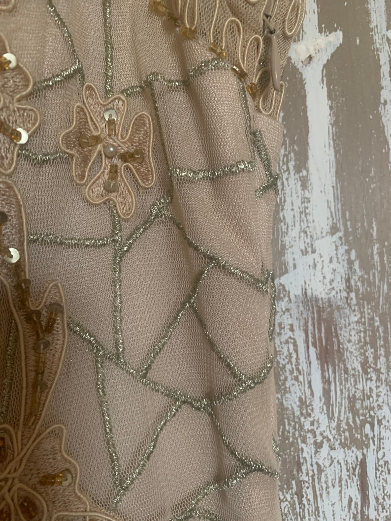 May include: Close-up of a beige fabric with intricate detailing. The fabric features a net-like pattern with silver thread accents. Floral and geometric designs are embellished with sequins and beads, creating a textured and elegant appearance. The overall aesthetic suggests formal wear.