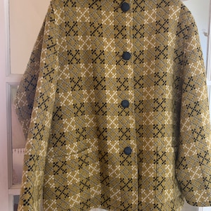 Op de afbeelding: Een vintage jas met capuchon, met een geometrisch patroon in olijfgroen, zwart en wit. De jas heeft een knoopsluiting met drie zwarte knopen en twee voorzakken. De jas hangt aan een zwarte hanger.