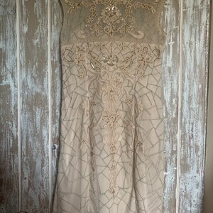 May include: A beige, sleeveless dress with intricate embroidery and sequin embellishments. The dress features a geometric pattern and a sheer neckline, creating a sophisticated and elegant look. The dress is hanging on a wooden door.