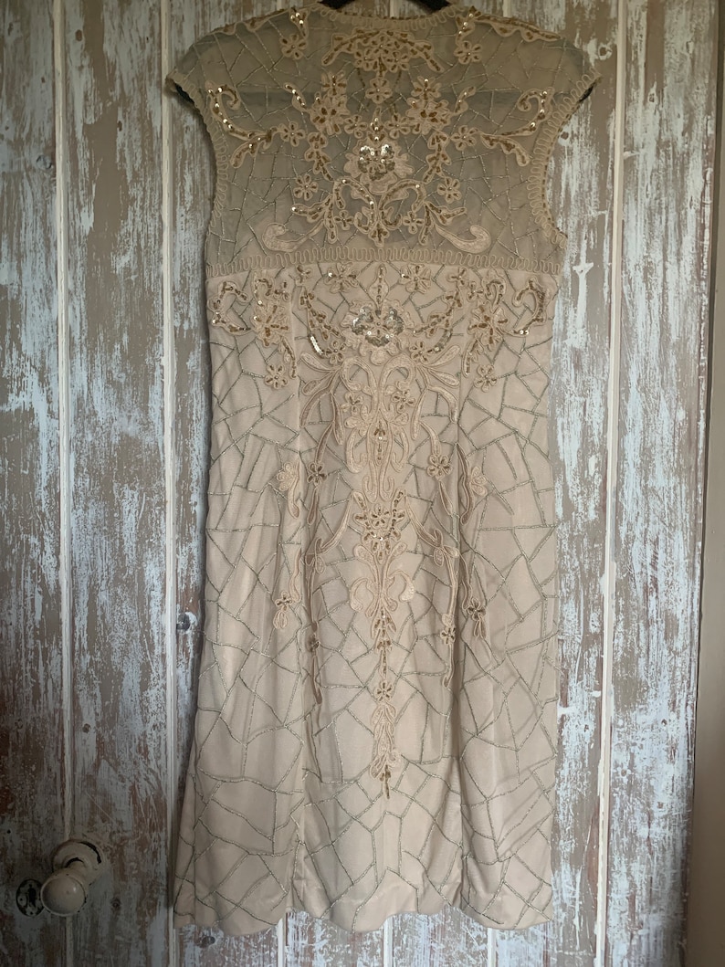May include: A beige, sleeveless dress with intricate embroidery and sequin embellishments. The dress features a geometric pattern and a sheer neckline, creating a sophisticated and elegant look. The dress is hanging on a wooden door.