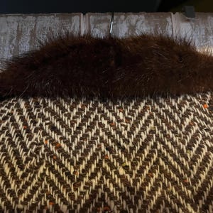 May include: Close-up of a brown and white herringbone pattern fabric with a dark brown fur collar. The fabric has flecks of orange and brown. The fur collar is thick and plush, adding a touch of luxury to the garment.