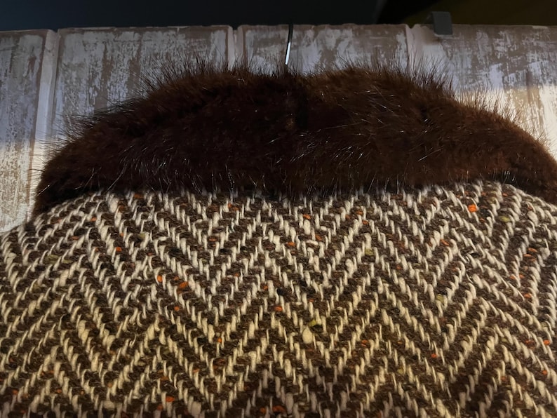 May include: Close-up of a brown and white herringbone pattern fabric with a dark brown fur collar. The fabric has flecks of orange and brown. The fur collar is thick and plush, adding a touch of luxury to the garment.