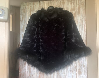 Puede incluir: Un poncho negro de piel sintética con un patrón texturizado y un cuello y ribete de piel. El poncho está colgado de una percha negra contra una puerta de madera de color claro.