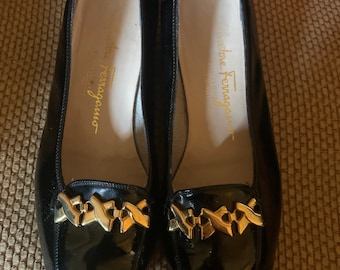 Puede incluir: Un par de mocasines de cuero negro con un acabado brillante. Los zapatos presentan un detalle de cadena dorada en la pala y el nombre de la marca "Salvatore Ferragamo" en letras doradas en el interior.