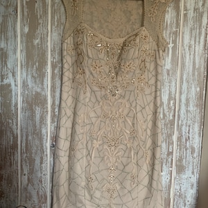 May include: A beige, knee-length dress with a square neckline and cap sleeves. The dress is adorned with intricate embroidery and beadwork in a floral and geometric pattern. The dress is hanging on a black hanger.