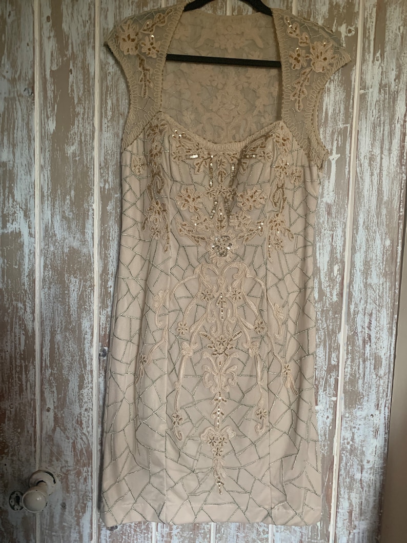 May include: A beige, knee-length dress with a square neckline and cap sleeves. The dress is adorned with intricate embroidery and beadwork in a floral and geometric pattern. The dress is hanging on a black hanger.