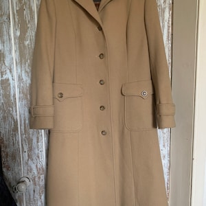 May include: A tan-colored, vintage overcoat with a classic design. The coat features a notched collar, button-down closure, two front pockets, and decorative buttons on the sleeves. The label reads "alexon Made in St. Britain".