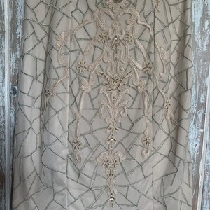 May include: A beige dress with intricate embroidery and sequin embellishments. The dress features a geometric pattern with silver stitching and floral and scrollwork designs. The bodice is adorned with sequins, adding a touch of sparkle to the elegant garment.