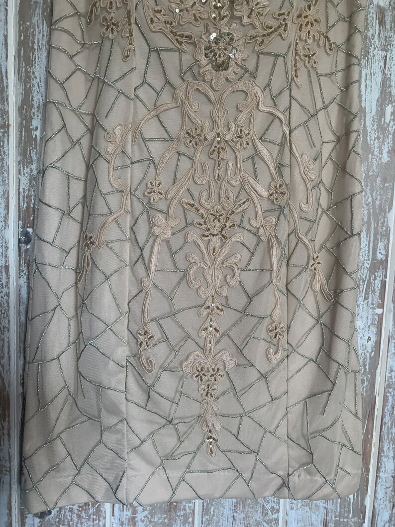 May include: A beige dress with intricate embroidery and sequin embellishments. The dress features a geometric pattern with silver stitching and floral and scrollwork designs. The bodice is adorned with sequins, adding a touch of sparkle to the elegant garment.