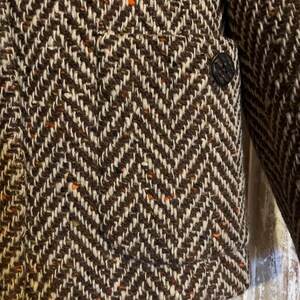 May include: Close-up of a brown and white herringbone tweed jacket. The jacket features a classic chevron pattern with flecks of orange. A single dark button is visible on the front, and a portion of the sleeve is also in view.
