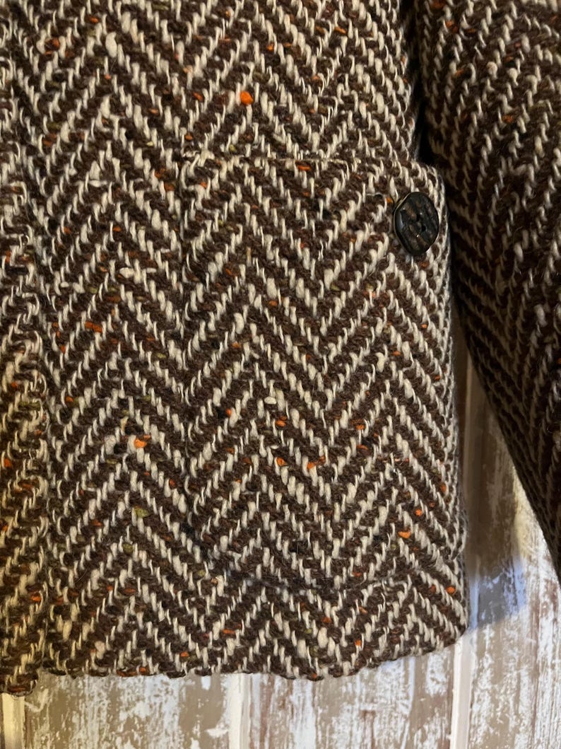 May include: Close-up of a brown and white herringbone tweed jacket. The jacket features a classic chevron pattern with flecks of orange. A single dark button is visible on the front, and a portion of the sleeve is also in view.