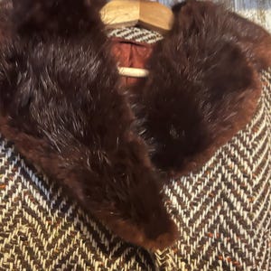 May include: A vintage brown and white herringbone pattern coat with a dark brown fur collar. The coat is hanging on a wooden hanger. The coat's interior is a reddish-brown color.
