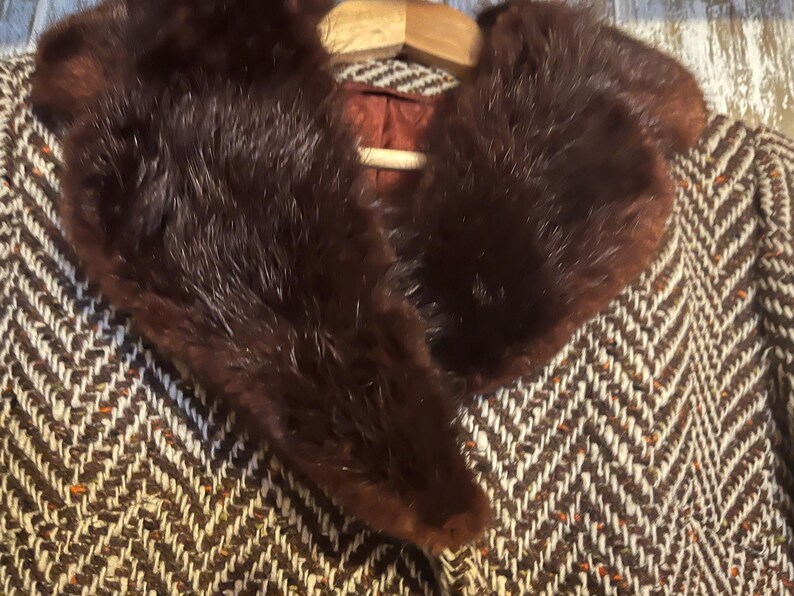 May include: A vintage brown and white herringbone pattern coat with a dark brown fur collar. The coat is hanging on a wooden hanger. The coat's interior is a reddish-brown color.