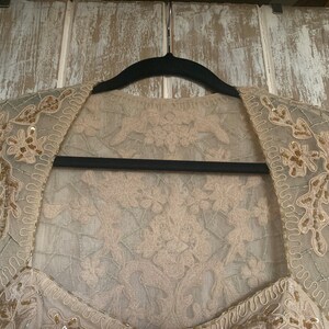 May include: A beige, embellished bolero jacket with floral and sequin details. The jacket is made of a sheer fabric and is hanging on a black hanger against a light-colored wooden background. The bolero has a square neckline.