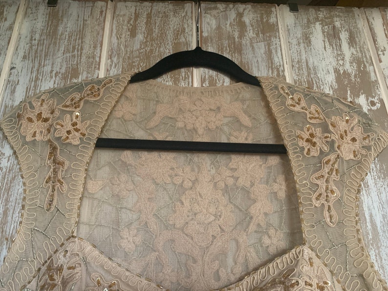 May include: A beige, embellished bolero jacket with floral and sequin details. The jacket is made of a sheer fabric and is hanging on a black hanger against a light-colored wooden background. The bolero has a square neckline.