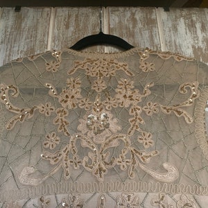 May include: A beige sleeveless top with intricate floral and geometric embroidery. The delicate design features sequins and beadwork, creating a shimmering effect. The top is hanging on a black hanger against a light-colored wooden background.