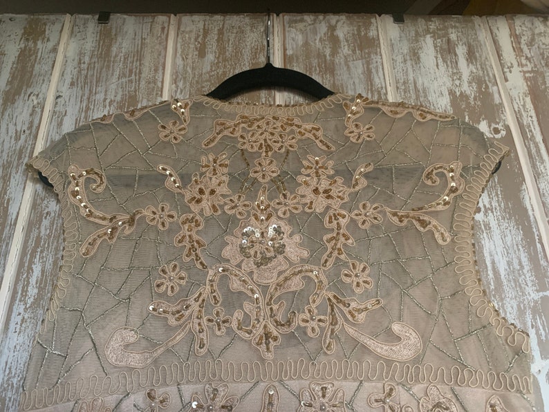 May include: A beige sleeveless top with intricate floral and geometric embroidery. The delicate design features sequins and beadwork, creating a shimmering effect. The top is hanging on a black hanger against a light-colored wooden background.
