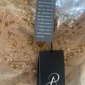May include: Close-up of a beige dress with intricate floral embroidery and beadwork. The dress has a delicate, sheer overlay. A black tag with the Adrianna Papell logo and text about the design's craftsmanship hangs from the garment.