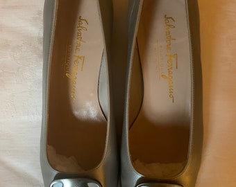 May include: A pair of silver-gray Salvatore Ferragamo Boutique pumps. The shoes have a rounded toe and a silver-tone buckle. The brand name is visible on the insole and the buckle. The shoes are displayed on a light-colored surface.