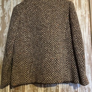 May include: A vintage jacket with a brown and cream herringbone pattern. The jacket has a dark brown fur collar. The jacket is displayed against a weathered wooden background.
