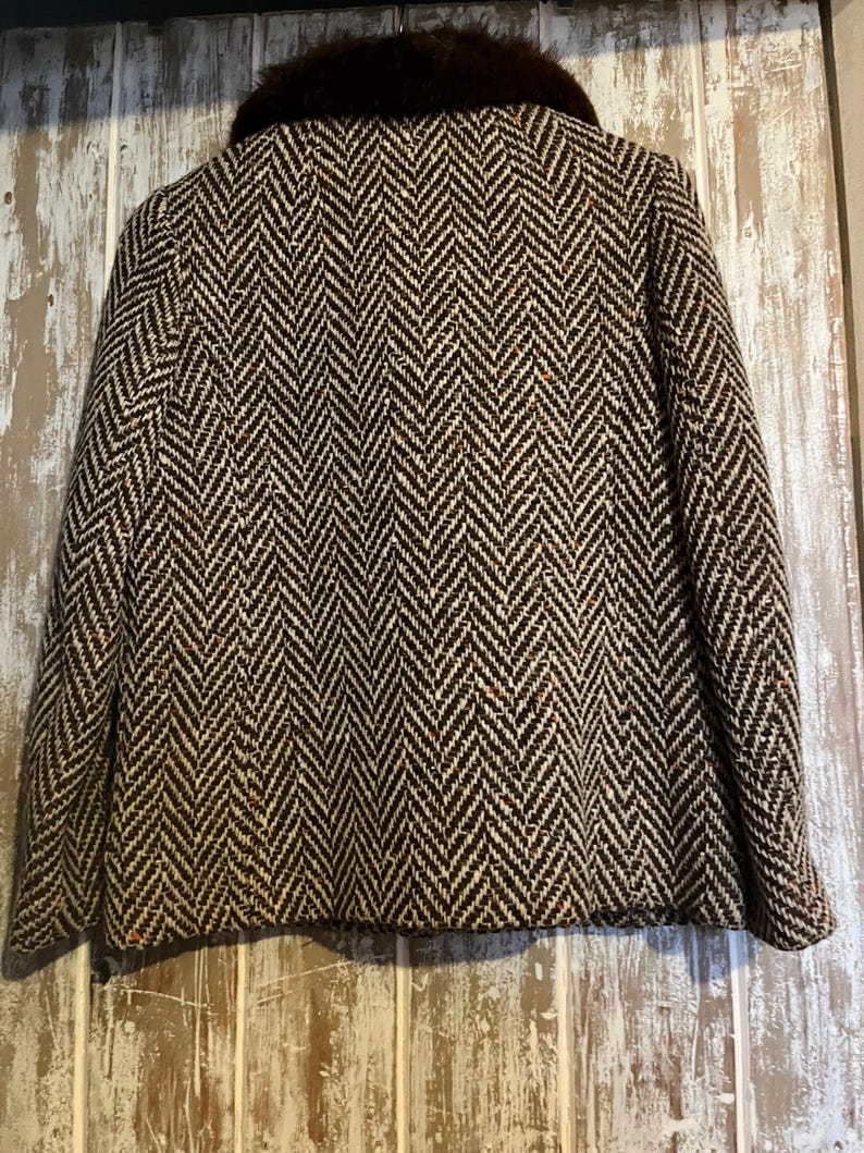 May include: A vintage jacket with a brown and cream herringbone pattern. The jacket has a dark brown fur collar. The jacket is displayed against a weathered wooden background.