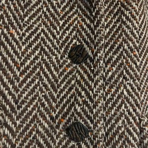 May include: Close-up of a brown and white herringbone tweed fabric with three dark, textured buttons. The fabric has a classic, woven pattern with flecks of orange and brown.