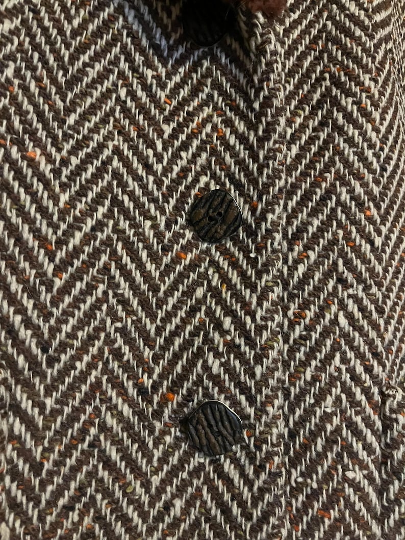 May include: Close-up of a brown and white herringbone tweed fabric with three dark, textured buttons. The fabric has a classic, woven pattern with flecks of orange and brown.