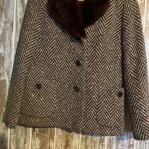 May include: A brown and white herringbone tweed jacket with a dark brown fur collar. The jacket has three buttons down the front and two pockets. The jacket is hanging on a wooden hanger.