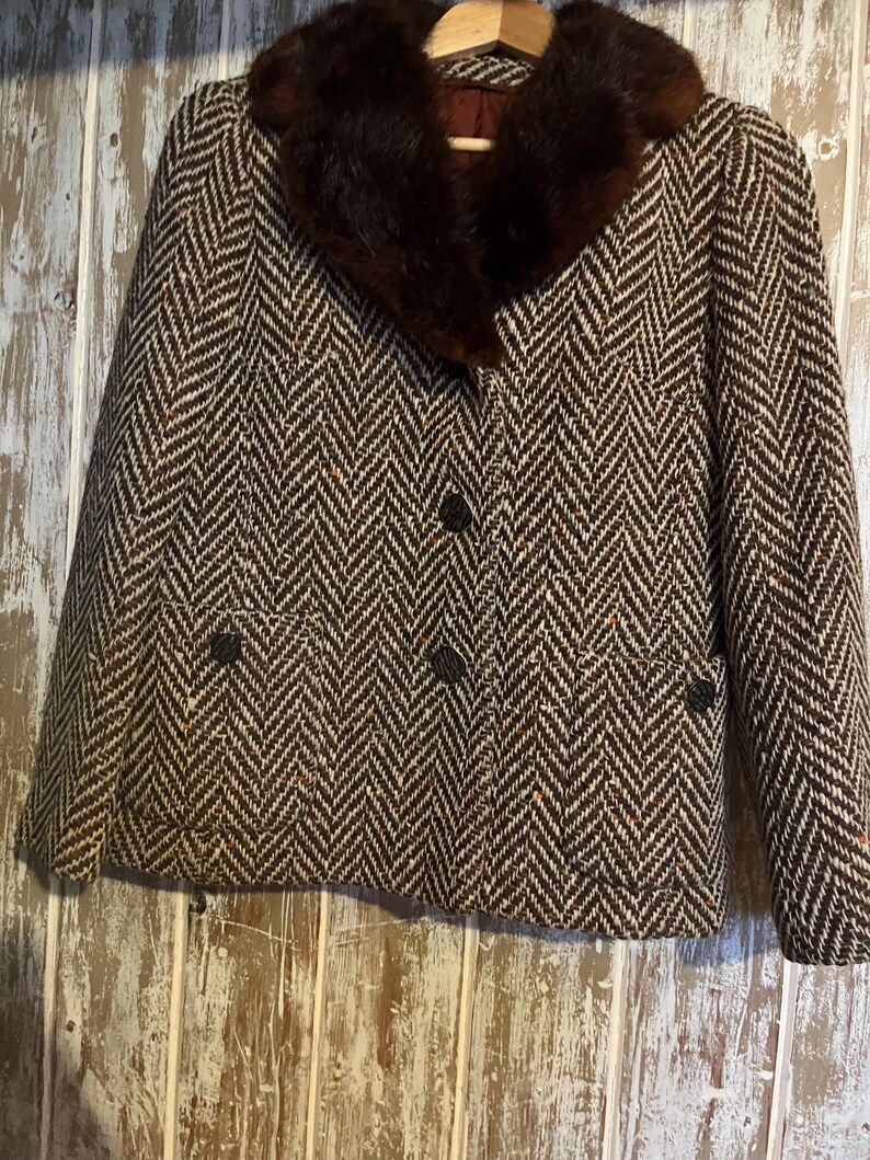 May include: A brown and white herringbone tweed jacket with a dark brown fur collar. The jacket has three buttons down the front and two pockets. The jacket is hanging on a wooden hanger.