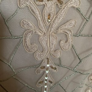 May include: Close-up of a fabric with an embroidered floral design. The embroidery is a light beige color, with silver thread accents and small sequins. The background is a sheer, light beige fabric.