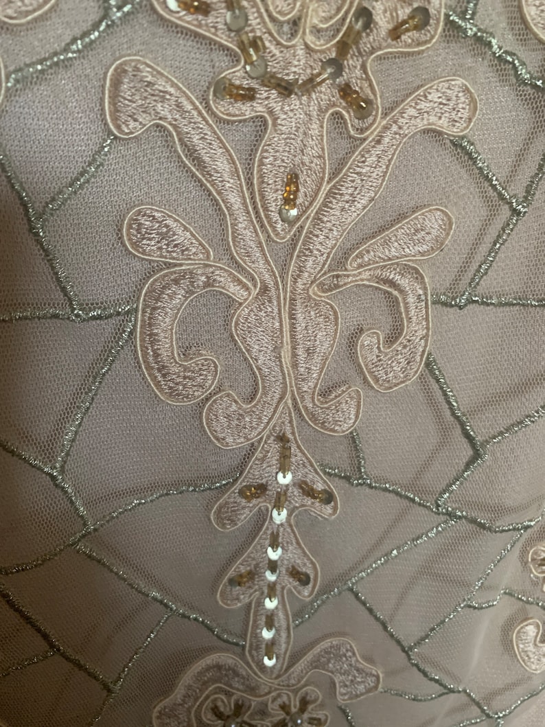 May include: Close-up of a fabric with an embroidered floral design. The embroidery is a light beige color, with silver thread accents and small sequins. The background is a sheer, light beige fabric.