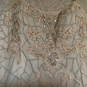 May include: A beige formal dress with intricate floral embroidery and sequin embellishments. The dress features a delicate, geometric net-like pattern and a scoop neckline. The design is detailed with gold and silver thread, creating a luxurious look.