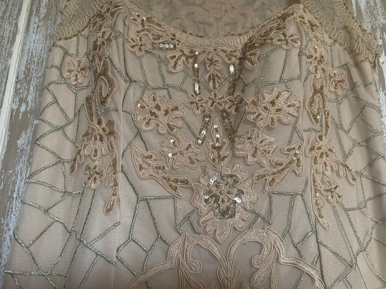 May include: A beige formal dress with intricate floral embroidery and sequin embellishments. The dress features a delicate, geometric net-like pattern and a scoop neckline. The design is detailed with gold and silver thread, creating a luxurious look.