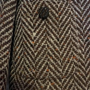 May include: Close-up of a brown and white herringbone tweed jacket. The jacket features a single, textured button and a small pocket. The fabric has flecks of orange throughout the weave, adding a subtle pop of color to the classic design.