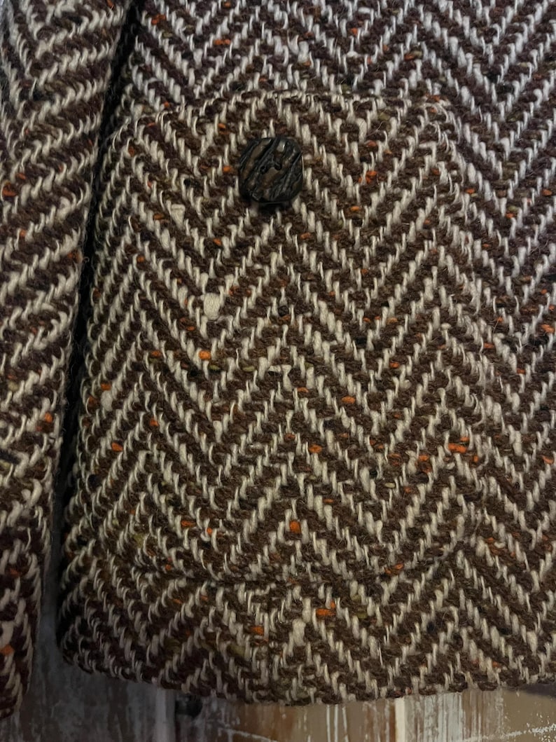 May include: Close-up of a brown and white herringbone tweed jacket. The jacket features a single, textured button and a small pocket. The fabric has flecks of orange throughout the weave, adding a subtle pop of color to the classic design.