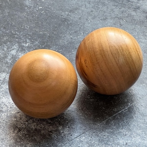 May include: Two wooden spheres with a warm, light brown color and visible wood grain. The spheres are smooth and round, resting on a gray, textured surface. The spheres appear to be made of a hardwood.