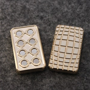 May include: Two rectangular, gold-colored metal objects. One has a series of eight circular silver magnets arranged in a pattern. The other has a grid pattern etched into its surface. Both objects have a polished finish.