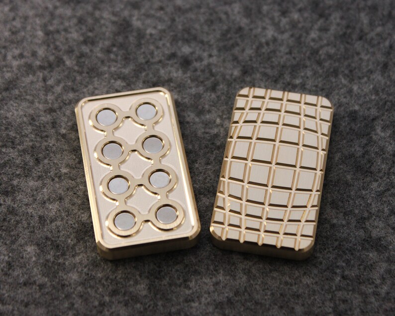 May include: Two rectangular, gold-colored metal objects. One has a series of eight circular silver magnets arranged in a pattern. The other has a grid pattern etched into its surface. Both objects have a polished finish.