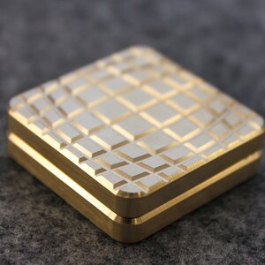 May include: A square, gold-colored metal box with a textured, diamond-patterned top. The box has a two-part design with a visible seam. The edges are smooth and the overall appearance is clean and modern.