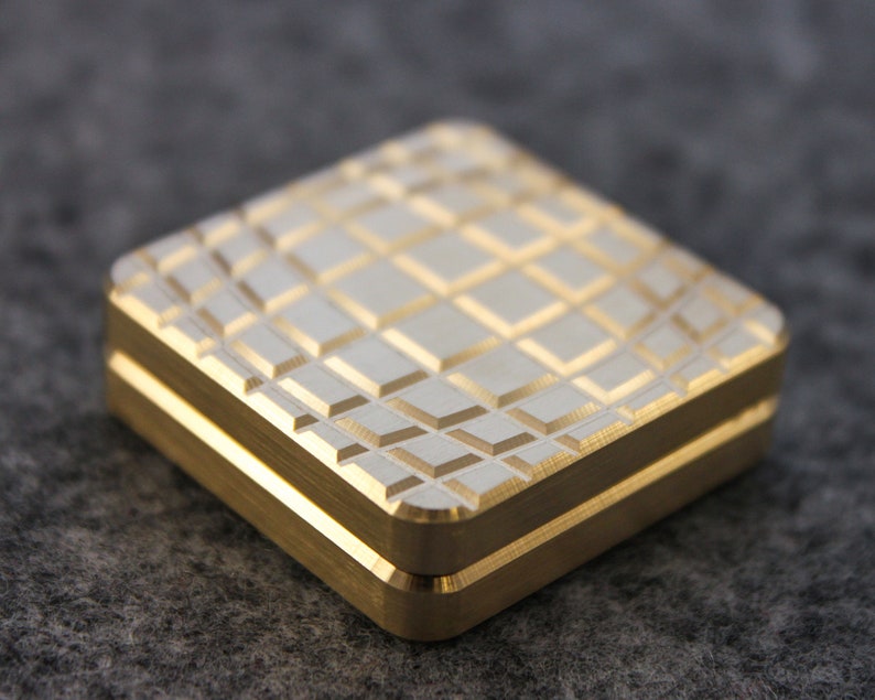 May include: A square, gold-colored metal box with a textured, diamond-patterned top. The box has a two-part design with a visible seam. The edges are smooth and the overall appearance is clean and modern.
